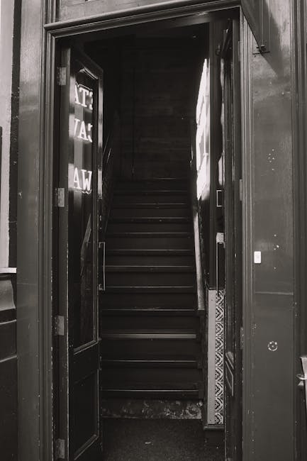 A narrow, steep staircase inside a property, featuring blue painted steps that are worn and chipped, revealing the underlying wood. The staircase is enclosed by white-painted walls, with some areas sh