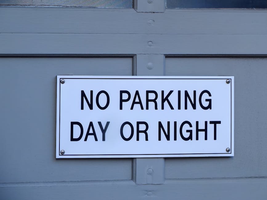 A rectangular white metal sign with black uppercase text reading 'NO PARKING DAY OR NIGHT' is fixed to the light grey exterior wall of a building, utilizing four screws, one at each corner. The sign i