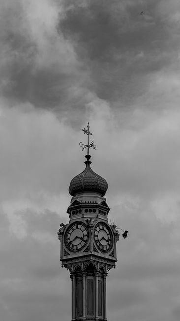 A black and white photograph of a historic clock tower featuring a rounded, domed roof topped with a weather vane, located in an urban setting. The tower's clock faces are visible, showing the time as