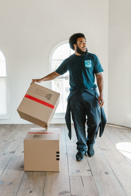 A person wearing a dark blue T-shirt is seen packing a cardboard box labeled 'Glass' with white packing peanuts inside a home. The individual is placing a clear glass bottle into the box, which is par